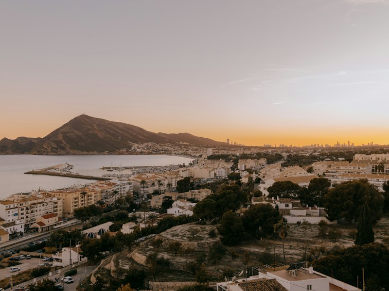 Panoramisch uitzicht op Altea in het gouden uur: huizen van de stad langs de baai, de jachthaven en de kustlijn, met de zee en de Sierra de Bernia op de achtergrond